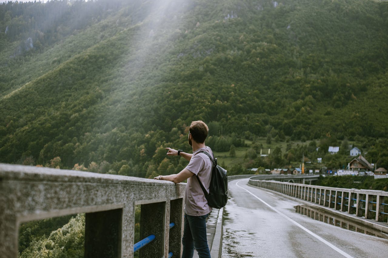 Caucasian man with backpack points across a bridge at lush mountains on a sunny day.