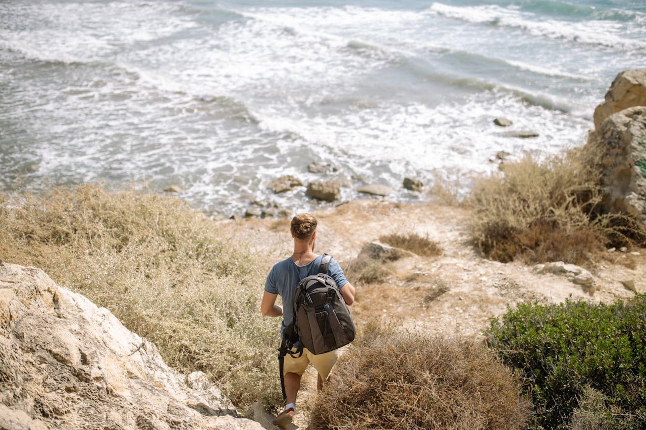 A solo backpacker walking down a rocky path towards the ocean, surrounded by coastal vegetation.