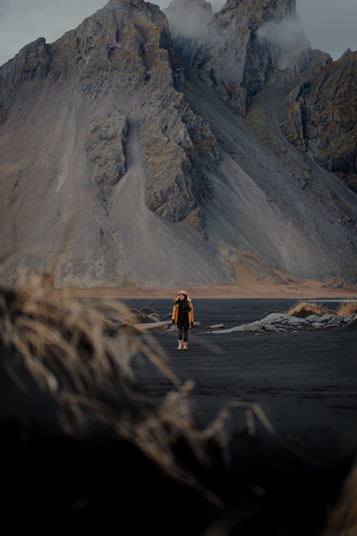 A solitary figure in a yellow jacket hikes on dark terrain with rugged mountains in the background.